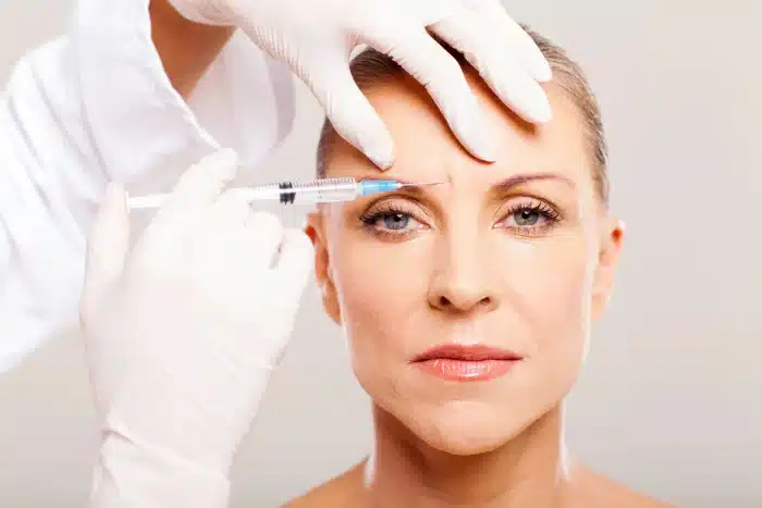 A person with a neutral expression receives a cosmetic injection in the forehead from a gloved professional. The individual has light skin and is framed from the chest up, with their hair pulled back, while the medical professional’s gloved hands are visible administering the injection.