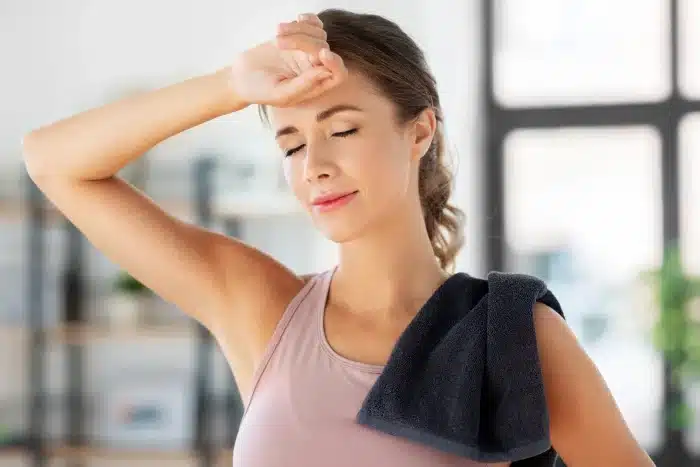 A woman in athletic wear rests indoors with a towel draped over her shoulder, wiping her forehead with her hand and eyes closed, appearing relaxed and refreshed. A bright and softly blurred background suggests a home or gym setting.