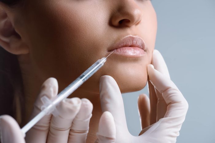 A close-up of a person receiving a lip injection. The person's face is partially visible with emphasis on their lips. A gloved hand is holding a syringe and administering the injection to the lower lip. The background is a neutral, light blue color.