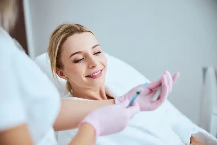 A smiling woman with blonde hair reclines on a white medical bed while a healthcare professional wearing pink gloves prepares a syringe. The setting appears to be a clinical environment with soft lighting, indicating a possible medical or cosmetic procedure.