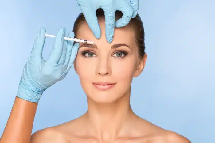 A woman with a serene expression is receiving a cosmetic injection in her forehead. She has her hair tied back and is against a blue background. The procedure is done by a person wearing blue medical gloves.