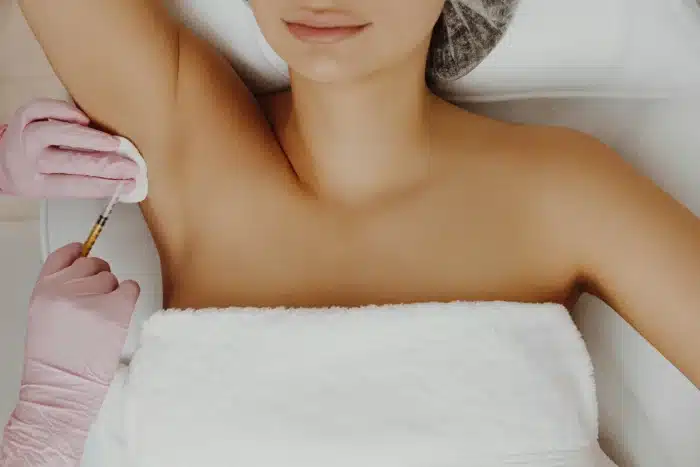 A woman lies on a spa bed with a white towel covering her chest. She has a neutral expression, and her right arm is raised. A practitioner's hand, wearing pink gloves, is applying an injection to her underarm area using a syringe.