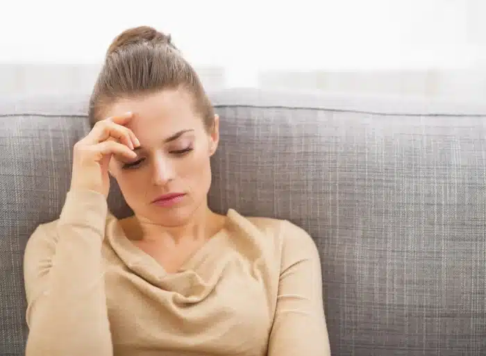 A woman with light brown hair in a bun sits on a gray couch, looking down with a hand on her forehead. She appears to be deep in thought or worried, wearing a beige long-sleeve top against a blurred light background.