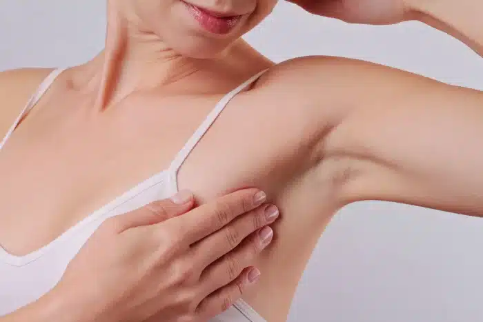 A person wearing a white tank top is shown with their right arm raised, using their left hand to check their right underarm area. They are performing a self-examination, possibly for breast health or skin condition. The background is plain and light-colored.