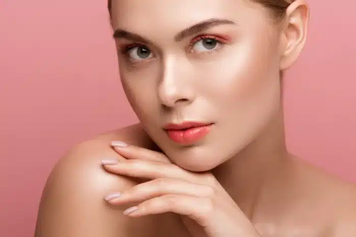 A woman with natural makeup and dewy skin, accentuated by enhanced dermal fillers, poses in front of a pink background. She rests her chin on her hand, showcasing neatly manicured nails.