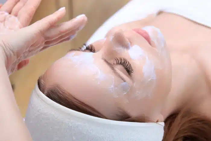 A person lying on their back receives a facial treatment. Cream is applied to their face by a pair of hands. They have their eyes closed, are wearing a headband, and have long eyelashes. The background is out of focus, highlighting the serene setting.