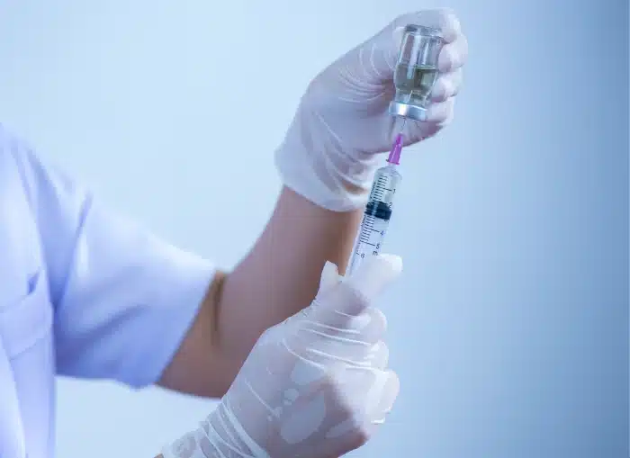 A healthcare professional wearing gloves is filling a syringe with liquid from a vial. The background is blurred, with a blue tint. The person's clothing is a light color, possibly a uniform.