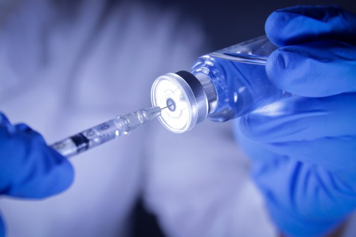 A person wearing blue gloves holds a vial and draws a liquid with a syringe. The scene suggests a medical or clinical setting.