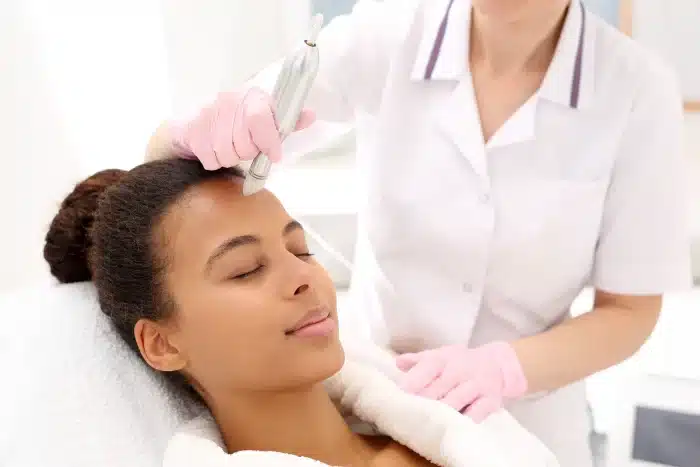 A person with a relaxed expression is lying down with their eyes closed while another individual, wearing a white uniform and pink gloves, is holding a skincare device and performing a facial treatment on their forehead.