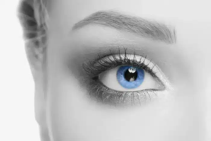 Close-up of a person's face, focusing on their bright blue eye and eyebrow. The image is in grayscale except for the eye, which is vividly blue. The eyelashes are long and defined, and there's light makeup around the eye area.