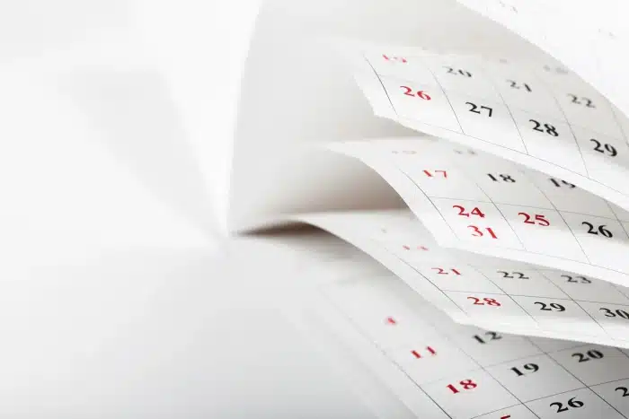 Close-up of a calendar with several pages fanned out, displaying dates in a grid format. The dates are numbered in red and black, with the weekends commonly highlighted in red. The background is minimalistic and white, giving a clean and organized impression.