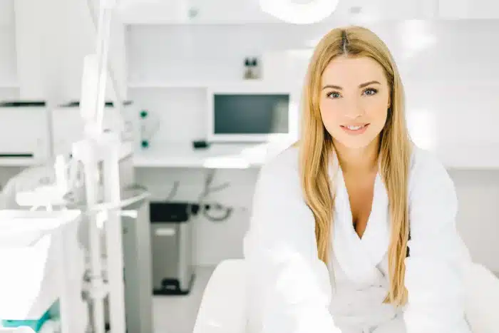 A woman with long blonde hair, wearing a white coat, is seated in a bright, modern medical or dental office. She is smiling at the camera. The background includes medical equipment and a computer.