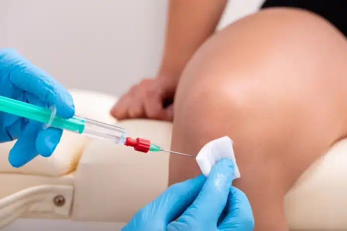 A healthcare professional wearing blue gloves prepares to administer an injection to a patient's knee. The professional holds a syringe and a piece of cotton, while the patient sits on an examination table.