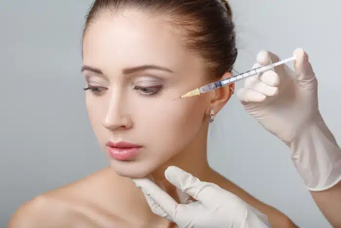 A close-up of a woman's face receiving a cosmetic injection. A person wearing white gloves is holding a syringe and injecting it into the skin near the woman's eye. The woman appears calm and is looking slightly downward. The background is plain and light gray.