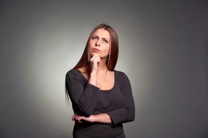 A woman with long brown hair and wearing a black dress is standing against a gray background. She has a thoughtful expression, with one hand resting on her chin and the other arm crossed over her body.