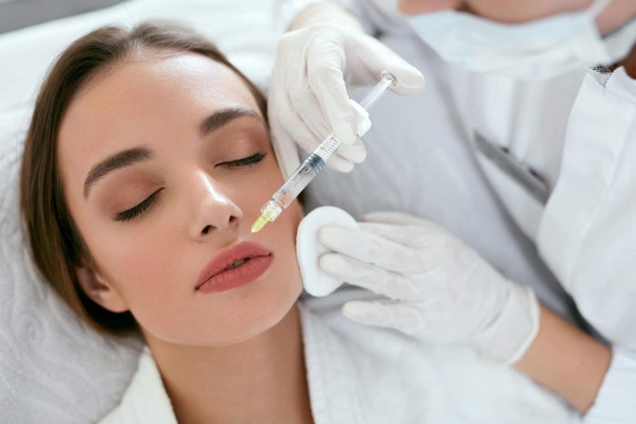A woman lying down with her eyes closed, receiving a cosmetic injection in her upper lip from a medical professional wearing gloves and holding a syringe. The procedure is being performed in a clinical setting.