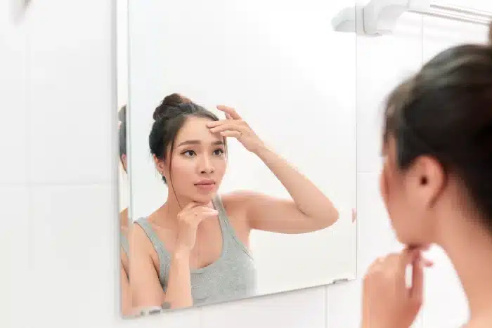 A woman with her hair in a bun is standing in front of a mirror, examining her face. She is wearing a gray tank top and touching her forehead with one hand while resting her chin on the other, perhaps contemplating Juvederm for acne scars. The background features white tiles and a towel rack.