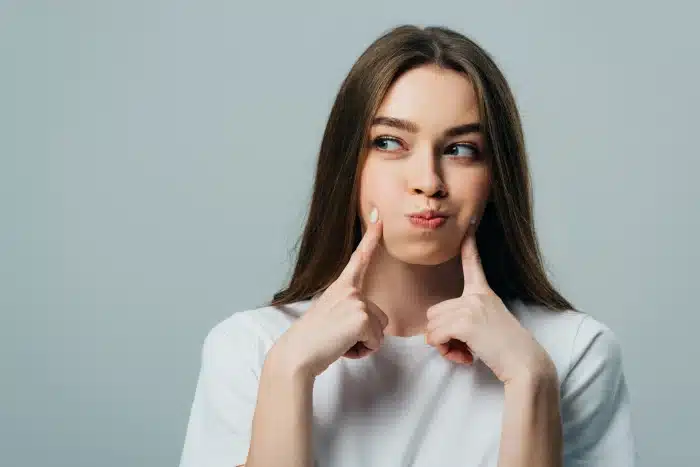A young woman with long brown hair wearing a white t-shirt is standing against a gray background. She is playfully pressing her cheeks with both index fingers and looking to the side with a puckered expression.
