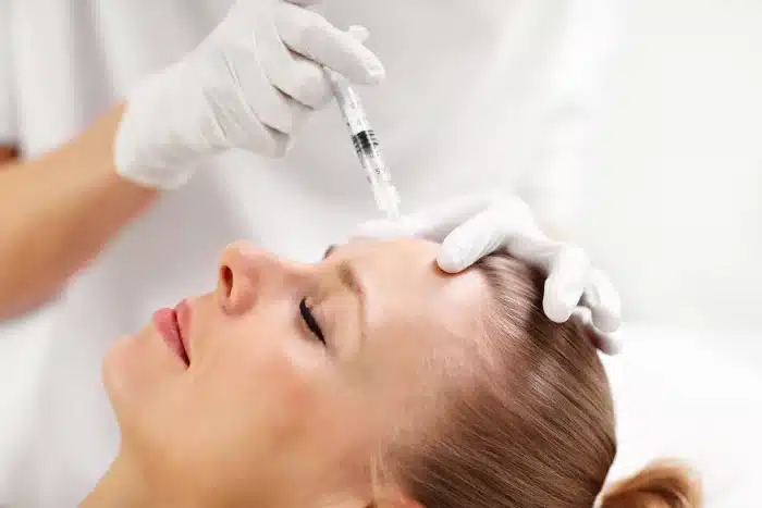 A person with closed eyes is receiving an injection in the forehead from another individual wearing white gloves. The scene appears to be a medical or cosmetic procedure, likely in a clinical or spa setting.