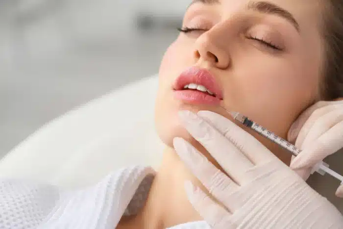 A woman with pink lips and closed eyes is receiving a lip injection. The procedure is being performed by a professional wearing white gloves, who is holding a syringe near the woman's lips. She is lying back and appears relaxed.