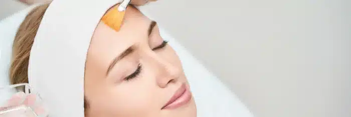 A person is lying down with a relaxed expression, wearing a white headband. A hand is applying a facial treatment with a brush to the person's forehead. The background is a neutral, light color.