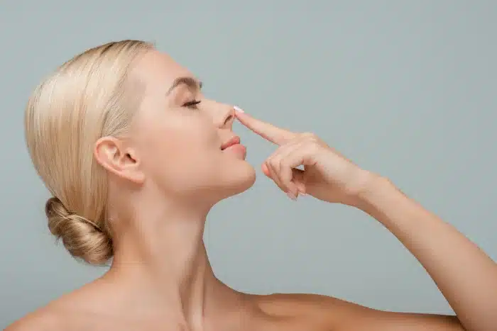 A woman with blonde hair pulled back in a bun is seen in profile against a neutral background. Her eyes are closed, and she has a serene expression as she gently touches the tip of her nose with one finger.