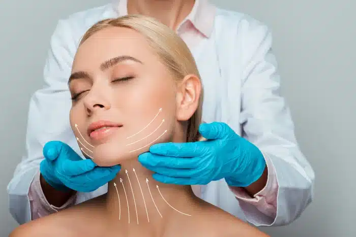 A person with closed eyes is sitting relaxed, while a professional wearing a white coat and blue gloves gently touches their face. White lines indicating potential lifting points are drawn on the person's cheek and chin area, suggesting a cosmetic procedure.