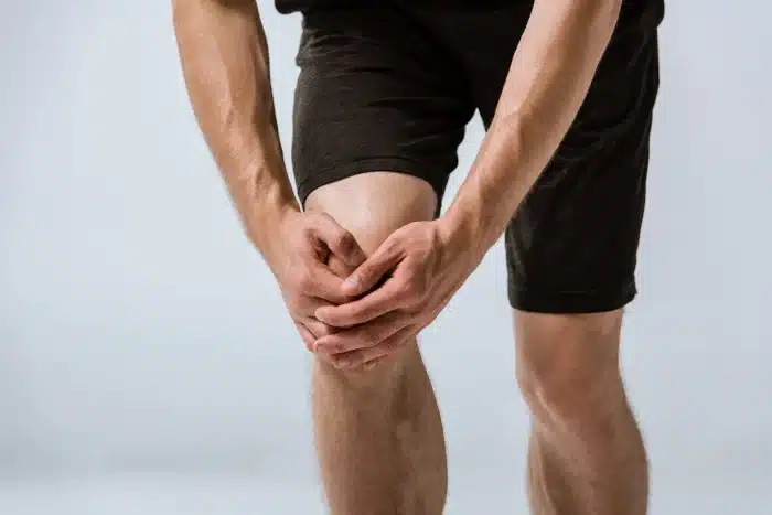 A person standing and holding their knee with both hands, possibly indicating pain or discomfort. The individual is wearing dark shorts and a dark shirt, and the background is plain and light-colored.