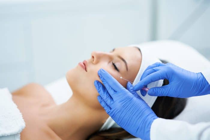 A close-up of a person with closed eyes receiving a cosmetic face treatment. They are lying down and have a white headband on. A practitioner wearing blue latex gloves is inserting a thin, hollow needle into the person's cheek. The background is blurred.
