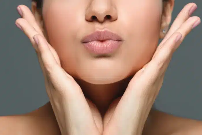 Close-up of a person with smooth, clear skin pouting their lips. Their hands are placed on either side of their face, gently cupping their cheeks. The background is a neutral grey.