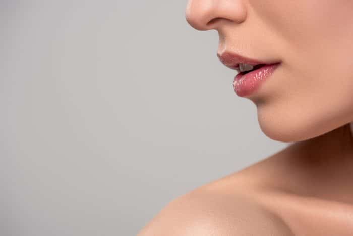Close-up of the lower half of a woman's face, focusing on her lips and chin. She has smooth skin and is wearing light pink glossy lipstick. The background is a solid, neutral gray. Her shoulder is slightly visible at the bottom of the image.