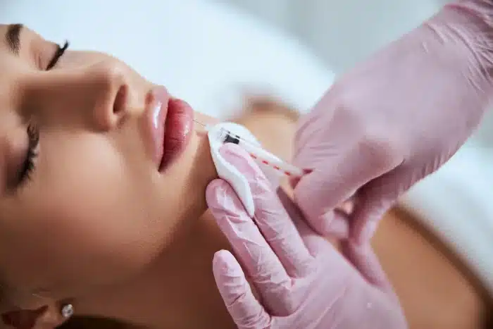 A person receiving a lip injection from a professional wearing pink gloves. The person has their eyes closed and a white cloth is held under their lower lip. The background is blurred, focusing attention on the procedure.