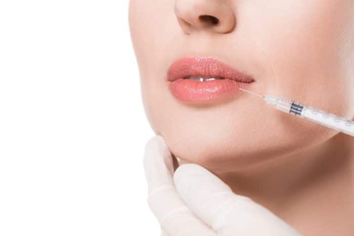A close-up of a woman's face with a syringe injecting a substance into her lips. She has light skin, pink lipstick, and is being administered the injection by a person wearing white gloves. The background is white.