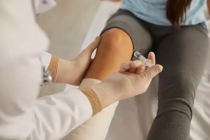 A medical professional wearing gloves administers an injection into the knee of a patient who has a bandaged lower leg. The patient is seated on a medical examination table, and the setting appears to be a clinical environment.