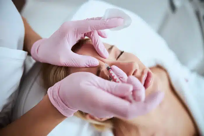 A person lies on a spa bed with eyes closed, receiving a facial treatment. A cosmetologist wearing pink gloves is gently touching the person's eyebrow with one hand while holding a cotton pad with the other. The setting appears to be a professional spa or clinic.