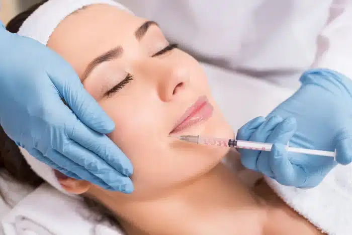 A relaxed woman is receiving a facial injection at a clinic. She is lying down with her eyes closed, and a practitioner, wearing blue gloves, is gently holding her face while administering the injection in the perioral area. The setting is clean and clinical.