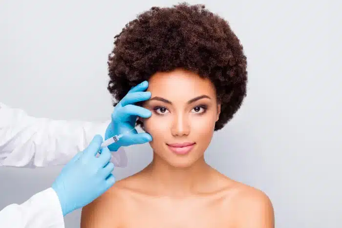 A person with natural curly hair is receiving a cosmetic injection in the face. A medical professional wearing blue gloves is holding the person's face with one hand and administering the injection with the other. The background is plain and light-colored.