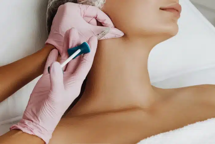Close-up of a person receiving a cosmetic injection in the jawline area. The individual administering the injection is wearing pink gloves and holding a syringe and a small blue device. The patient is lying down and appears relaxed.