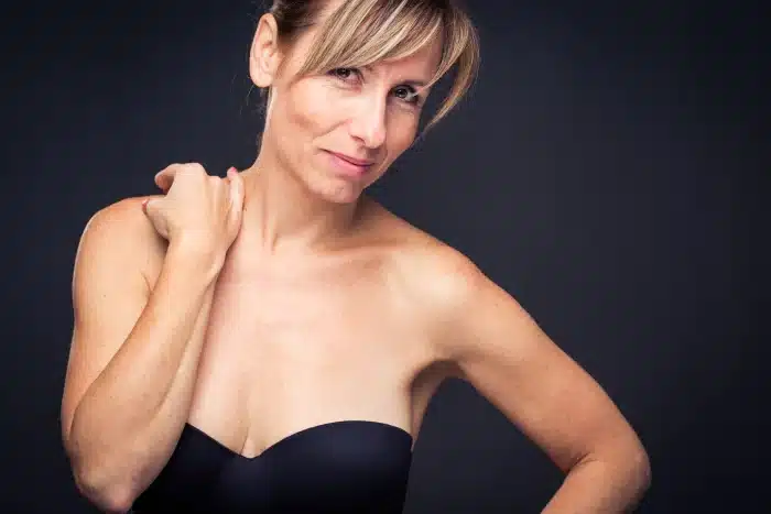A person with light skin and blonde hair in an updo is posing confidently against a dark background. They are wearing a black, strapless top and have one hand resting on their shoulder. The person is slightly smiling and looking directly at the camera.
