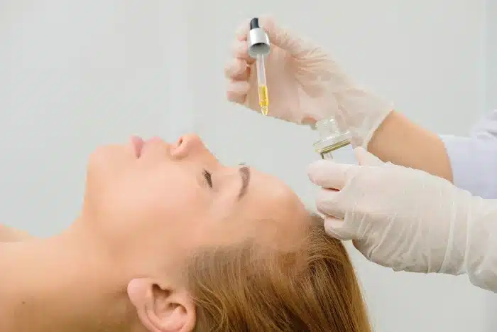 A person lies with eyes closed, receiving a facial treatment. Gloved hands apply an oil or serum from a dropper onto the person's skin. The background is out of focus, putting emphasis on the treatment and the person’s relaxed expression.