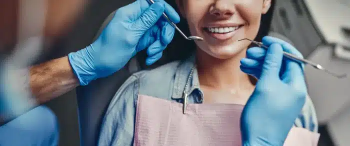 A patient wearing a dental bib is seated in a dentist's chair, smiling as a dentist in blue gloves examines their teeth using dental instruments.