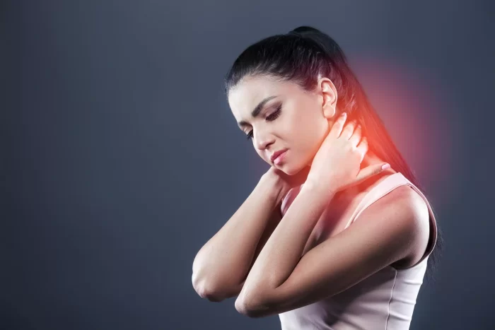 A woman with long dark hair tied back is holding her neck with both hands, wincing in pain. A red highlight emphasizes the area of discomfort, suggesting neck pain or strain. She is wearing a sleeveless top with a neutral background.