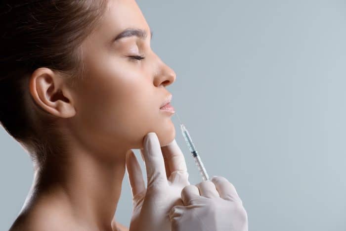 A person with closed eyes receives a cosmetic injection in the lips from a gloved professional. The background is plain and neutral, emphasizing the procedure.