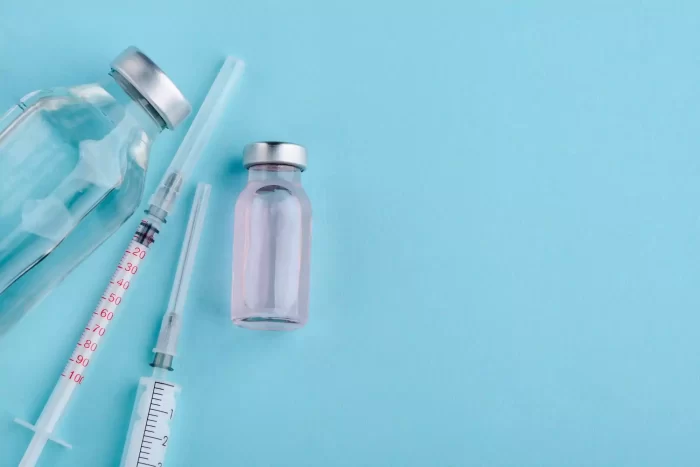 A flat lay of medical supplies including two vials, one filled with clear liquid and the other with a pink liquid, alongside a couple of syringes, all set against a light blue background.