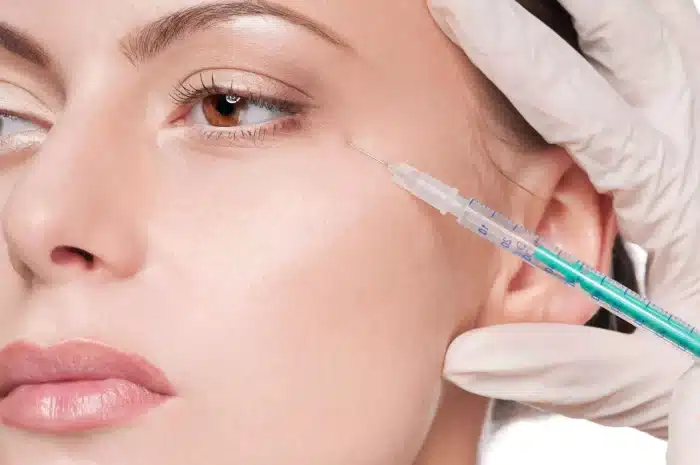 Close-up of a woman's face during a cosmetic procedure. A gloved hand is holding a syringe near the woman's eye, indicating that she is about to receive an injection, possibly for Botox or dermal filler treatment. The woman's skin looks smooth and clear.