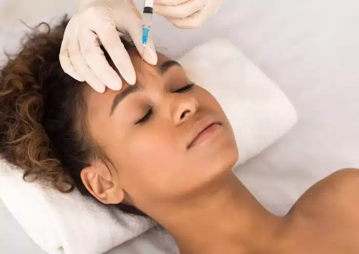 A person with eyes closed receives a facial injection from gloved hands. The individual is lying on a white towel, indicating a clinical or spa setting. Their hair is tied back, and the focus is on the syringe and the person's forehead.