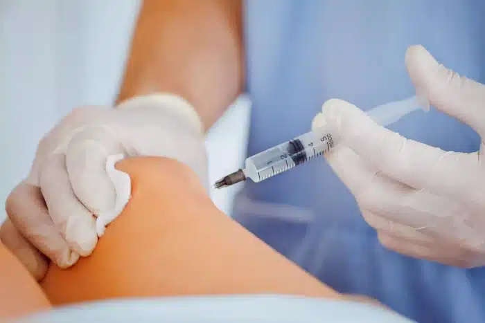 A medical professional in a blue uniform administers orthopedic injectables into a person's knee. Wearing gloves, they hold the knee steady with a cotton pad in one hand while directing the syringe with the other.
