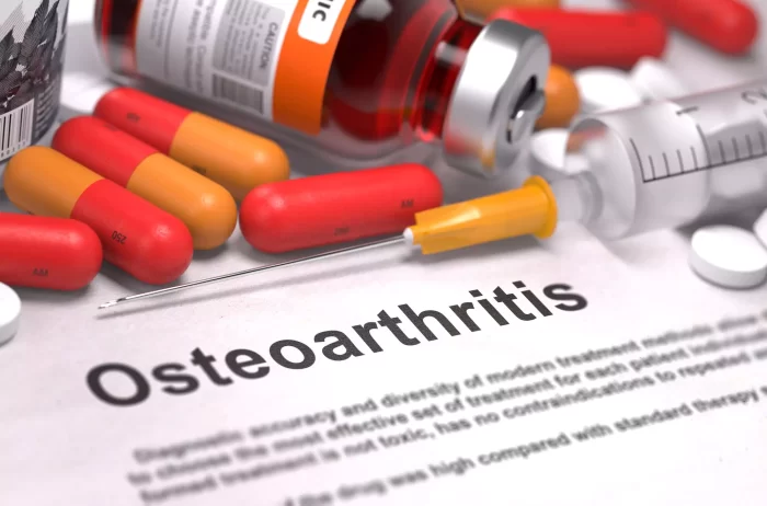 A document titled "Osteoarthritis" is surrounded by various medications, including red and yellow capsules, a syringe with a yellow cap, and a vial of liquid medicine. The setup suggests medical treatment or research related to osteoarthritis.