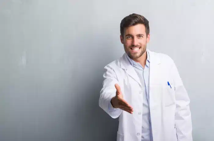 Smiling man in a white doctor's coat extends his right hand for a handshake against a plain, light-gray background. He has short brown hair, a beard, and is wearing a light blue shirt under the coat. A pen is visible in his coat pocket.