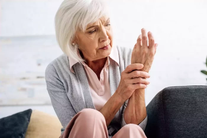 An elderly woman with short white hair sits on a sofa, looking down at her wrist with a concerned expression. She is wearing a light gray sweater over a pink blouse and appears to be examining or massaging her wrist, possibly in discomfort or pain.